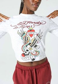 Valkoinen pitkähihaine crop-top, jossa on värikkäitä tatuointi-inspiroituja grafiikoita pääkallosta, kotkasta ja käärmeestä, sekä "Ed Hardy" -teksti ja punaisia korostuksia.