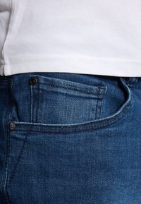 Jeans in denim blu scuro con vestibilità dritta classica, dotati di tasche anteriori e posteriori e dettagli di cucitura sottili. La texture è liscia.