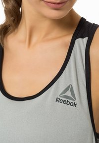 Jasnoszary sportowy tank top z czarnymi wykończeniami, z logo Reebok z lewej strony. Materiał wydaje się gładki i dopasowany.