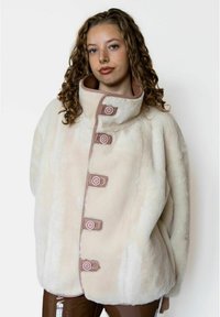 Nepbonten jas in crème met een hoge kraag, voorzien van vijf roze knopen en een zachte textuur. Het ontwerp is oversized met een gladde afwerking.