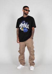 Sort grafisk t-shirt med farverigt design og tekst, kombineret med beige cargo-bukser med flere lommer og hvide sneakers.