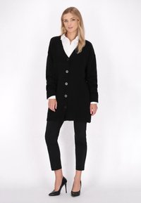 Schwarze Strickjacke mit V-Ausschnitt, ausgestattet mit sieben Knöpfen und gerippten Bündchen, kombiniert mit schwarzen, figurbetonten Hosen und High-Heels.