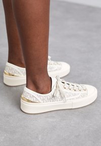 Witte canvas sneakers met gouden hardware accenten, met een gestructureerd logo patroon en een chunky zool, geregen met platte veters.