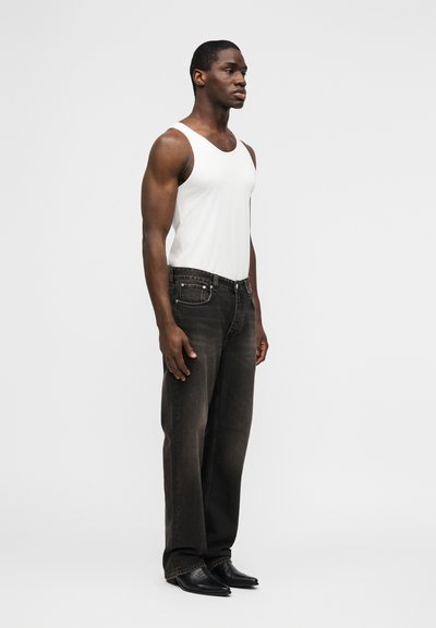 Άνδρας με λευκό αμάνικο μπλουζάκι, ξεθωριασμένο μαύρο τζιν και μαύρα παπούτσια, στέκεται μπροστά σε απλό λευκό φόντο, κοιτώντας προς τα δεξιά.