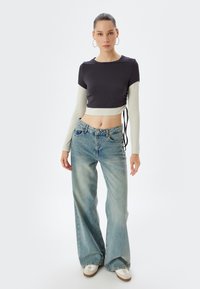 Afgemeten, aansluitend zwart topje met lange lichtgroene mouwen; hoog uitgesneden, losse blauwe denimjeans; witte en tan sneakers; minimalistisch ontwerp.