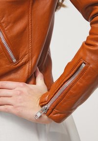 Brązowa skórzana kurtka o dopasowanym kroju, widoczne przeszycia oraz srebrne suwaki przy mankietach. Gładka tekstura z błyszczącym wykończeniem.