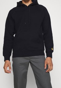 Persona che indossa una felpa nera con tasca frontale e logo dorato sulla manica, abbinata a pantaloni grigi, in piedi contro uno sfondo bianco.