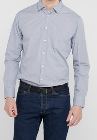 Hombre con una camisa de botones azul claro metida dentro de unos pantalones vaqueros azul oscuro, con un cinturón negro texturizado, de pie frente a un fondo liso.