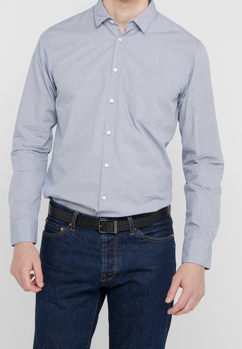Hombre con una camisa de botones azul claro metida dentro de unos pantalones vaqueros azul oscuro, con un cinturón negro texturizado, de pie frente a un fondo liso.