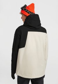 Personne portant une veste d'hiver noir et blanc, un bonnet orange vif et des lunettes de ski, de dos contre un fond uni.