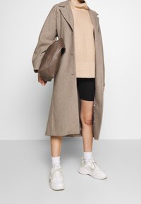 Lungo cappotto beige in lana con colletto a punte, abbinato a un maglione a coste marrone chiaro e pantaloni corti neri. Presenta scarpe da ginnastica bianche e una borsa marrone.