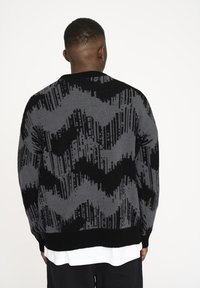 Grauer und schwarzer gestrickter Pullover mit Wellenmuster, gerippten Bündchen und Saum. Verfügt über einen Rundhalsausschnitt und ist mit einem weißen Hemd darunter kombiniert.