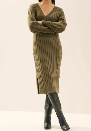 Femme en robe midi côtelée vert olive à manches longues avec col en V, portant des bottes noires montantes aux genoux, debout les bras croisés.
