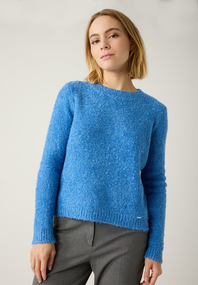Blauer flauschiger Pullover mit langen Ärmeln, Rundhalsausschnitt und gerippten Bündchen. Weiche Textur und lockere Passform, kombiniert mit grauen Hosen.