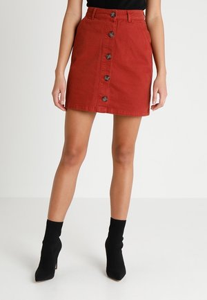 Femme portant une mini-jupe rouge taille haute à boutons sur le devant, associée à des bottines noires style chaussettes sur un fond uni.