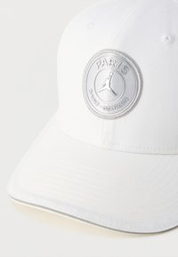 Λευκό καπέλο μπέιζμπολ με έναν στρογγυλό ασημένιο εμβληματικό δίσκο που φέρει τις λέξεις «PARIS» και «SAINT-GERMAIN» και σχέδιο σιλουέτας Jumpman. Ομαλή υφή υφάσματος.