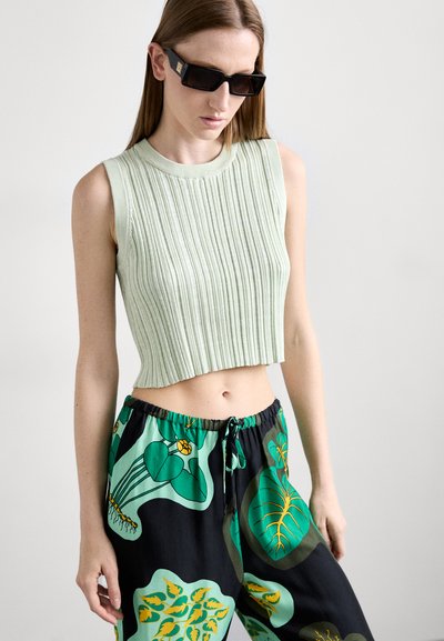 Top de tirantes cropped de color verde claro con textura acanalada, combinado con pantalones negros que presentan grandes patrones florales en verde y amarillo. Modelo usando gafas de sol.