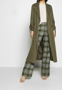 Cappotto lungo verde oliva con bottoni doppi, indossato sopra pantaloni a gamba larga grigi a scacchi con accenti gialli. Sandali beige completano il look.