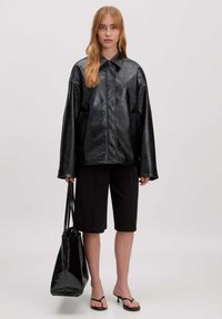 Zwarte faux leren jas met een kraag, oversized pasvorm en lange mouwen; gecombineerd met zwarte bermuda shorts en een glanzende zwarte handtas.