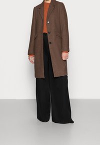 Person in einem braunen knöpfbaren Mantel über einem orangefarbenen Pullover und weit geschnittenen schwarzen Hosen, steht vor einem schlichten hellen Hintergrund.