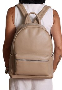 Persoon met een mouwloos wit topje en een beige leren rugzak met een voorzak met ritssluiting, zichtbare tatoeages op de armen.