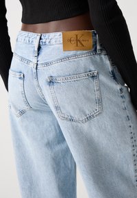 Lyseblå høy Midje jeans laget av denim, med en brun lærlapp med "CK"-logo, fem lommer og rette ben.