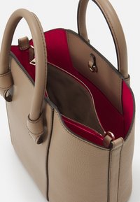 Beige nahkalaukku, jossa on rakenteellinen muotoilu, kaksinkertaiset kahvat, punainen sisustus ja vetoketjulli tasku turvalliseen säilytykseen. Huomattavat ompeluyksityiskohdat.