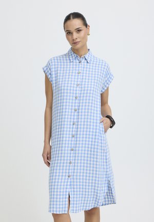 Naine, kes kannab lühikeste varrukatega, nööpidega ees sinise-valge ristikruvi mustriga kleiti kraega, poseerib ühe käega taskus heledal taustal.