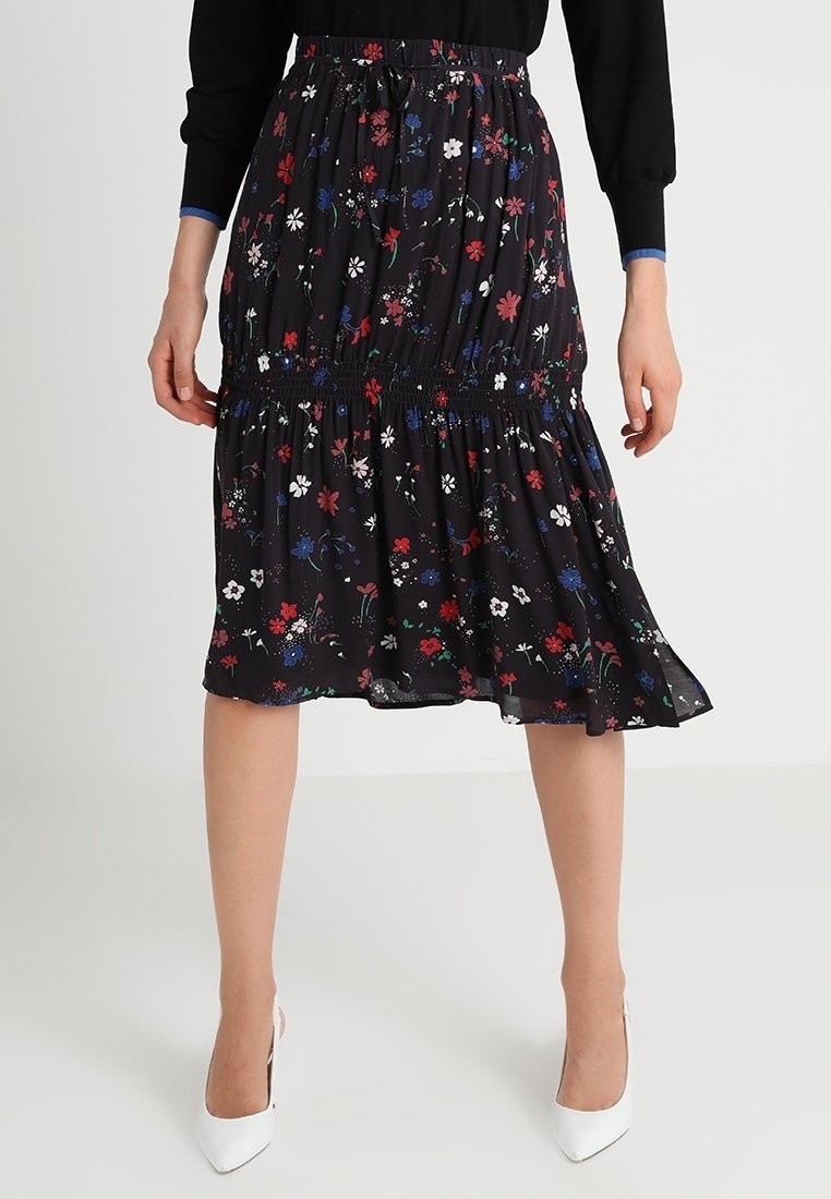 Jupe midi noire imprimé floral rouge, blanc et bleu, taille froncée et ourlet à volants, portée avec des talons blancs et un top noir à manches longues.