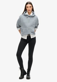 Grauer Reißverschluss-Hoodie mit großen Taschen, Kordelzugkapuze und weichem Material, über einem weißen Shirt getragen. Kombiniert mit schwarzen Skinny Jeans und Ankle Boots.