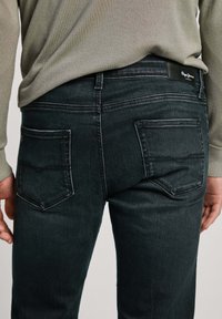 Person som bär svarta denimjeans med bakfickor och en ljus olivgrön tröja, sedd från midjan och bakifrån.