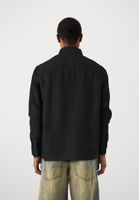 Camisa negra de manga larga con cuello, con una tela texturizada. La parte de atrás muestra puños abotonados y un dobladillo recto, combinada con jeans claros desgastados.