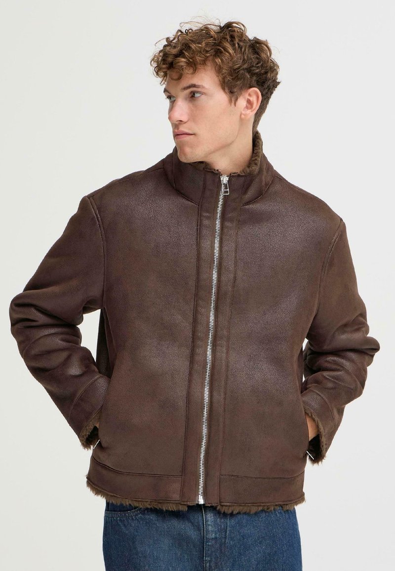 Braune Kunstlederjacke mit hohem Kragen, silbernem Reißverschluss und ausgefranstem Saum. Glatte Textur mit einem warmen Futter, getragen über Denim.