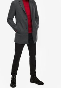 Cappotto in misto lana grigio con un solo bottone, revers a punta e tasche laterali, sovrapposto a un maglione rosso e abbinato a pantaloni neri e stivali con lacci.