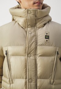 Kurtka puchowa w jasnobeżowym kolorze, z wysokim kołnierzem, dużym zapięciem na guziki oraz zapinanymi na zamek bocznymi kieszeniami. Tkanina o teksturze z pikowanym wzorem.