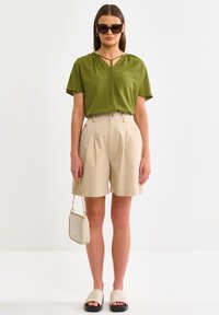 Alyvuogių žalia trumpų rankovių viršutinė dalis su rakto skylutės detale, derinama su šviesiai rudais klostuotais šortais. Papuošalai: grietinėlės spalvos per petį nešiojama rankinė ir juodi šlepečiai.