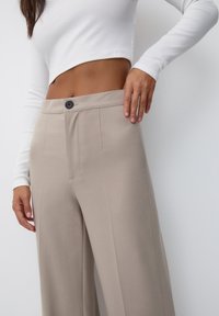 Žena v béžových širokých kalhotách a bílé dlouhé cropped topu, s jednou rukou opřenou o pás kalhot.