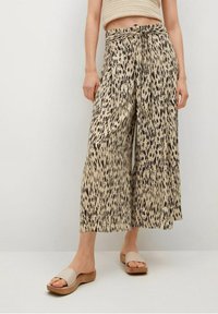 Culottes beige à jambes larges avec un motif animalier noir, dotées d'une ceinture à nouer et d'une texture lisse. Portées avec des sandales beiges plates.