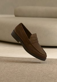 Καφέ μοκασίνι σουέντ με μαύρη σόλα που αιωρείται πάνω από μπεζ χαλί μπροστά από έναν ανοιχτό μπεζ καναπέ.