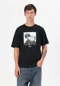 Camiseta de algodón negra con un corte recto, que presenta un gráfico en blanco y negro de dos individuos y una etiqueta de "oasis" en el centro inferior.