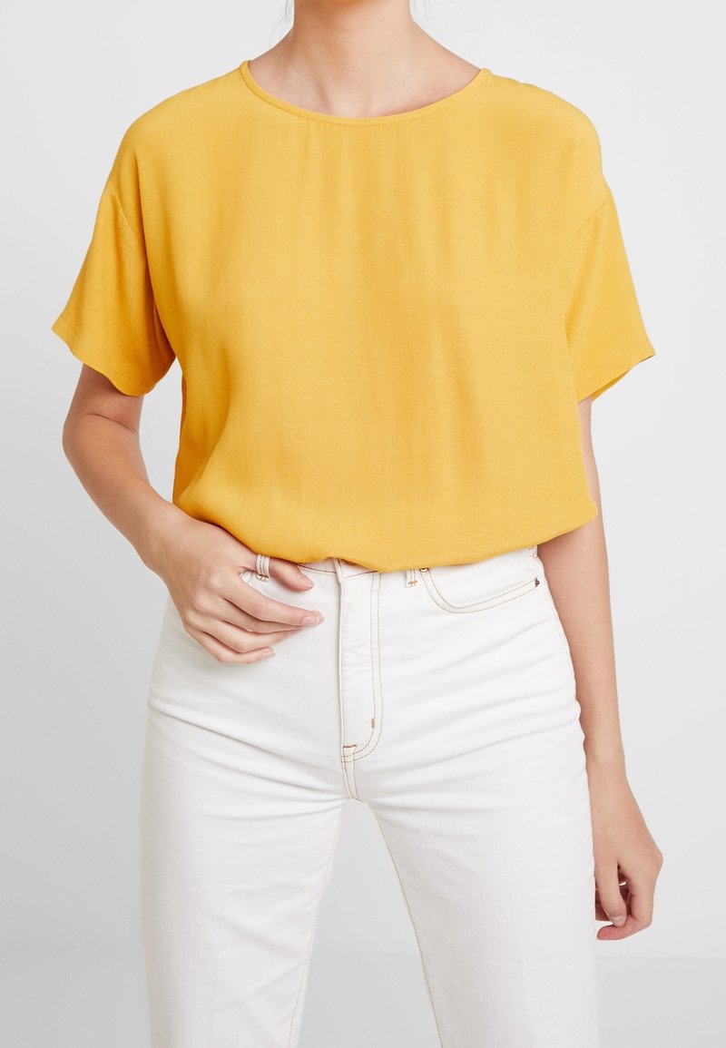 Blouse jaune à manches courtes avec un col rond, confectionnée dans un tissu léger, associée à un jean blanc taille haute. Texture lisse.
