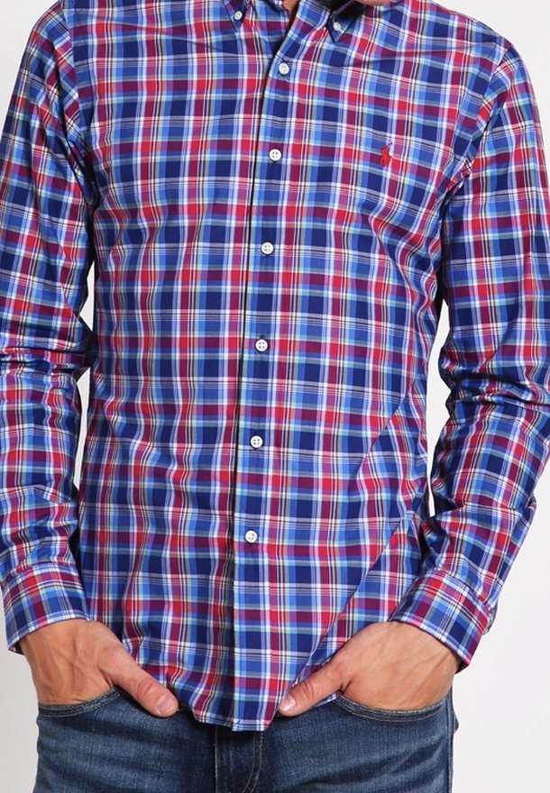 Homme portant une chemise à carreaux bleue, rouge et blanche à manches longues, avec les mains dans les poches.
