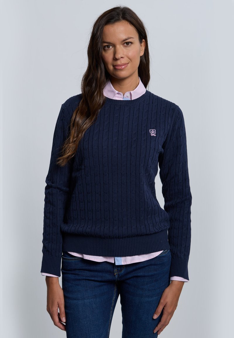 Suéter azul marino de punto con cable, con puños y dobladillo acanalados, usado sobre una camisa rosa con cuello. Presenta un pequeño logo en el lado superior izquierdo.