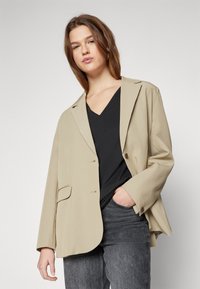Blazer beige su misura con rever e chiusura a bottone singolo. Indossato sopra un top nero a V, abbinato a jeans grigio scuro. Texture morbida.