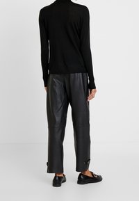 Schwarzer, langärmliger Strickpullover kombiniert mit schwarzen Lederhosen. Die Hosen haben eine lockere Passform und Knopfdetails an den Bündchen.