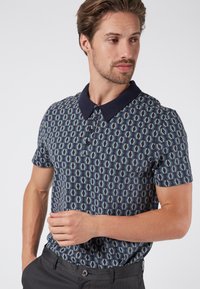 Kortärmad polo skjorta med ett geometriskt mönster i blått och beige, med en marinblå krage och plagg med tre knappar. Slät tygtextur.