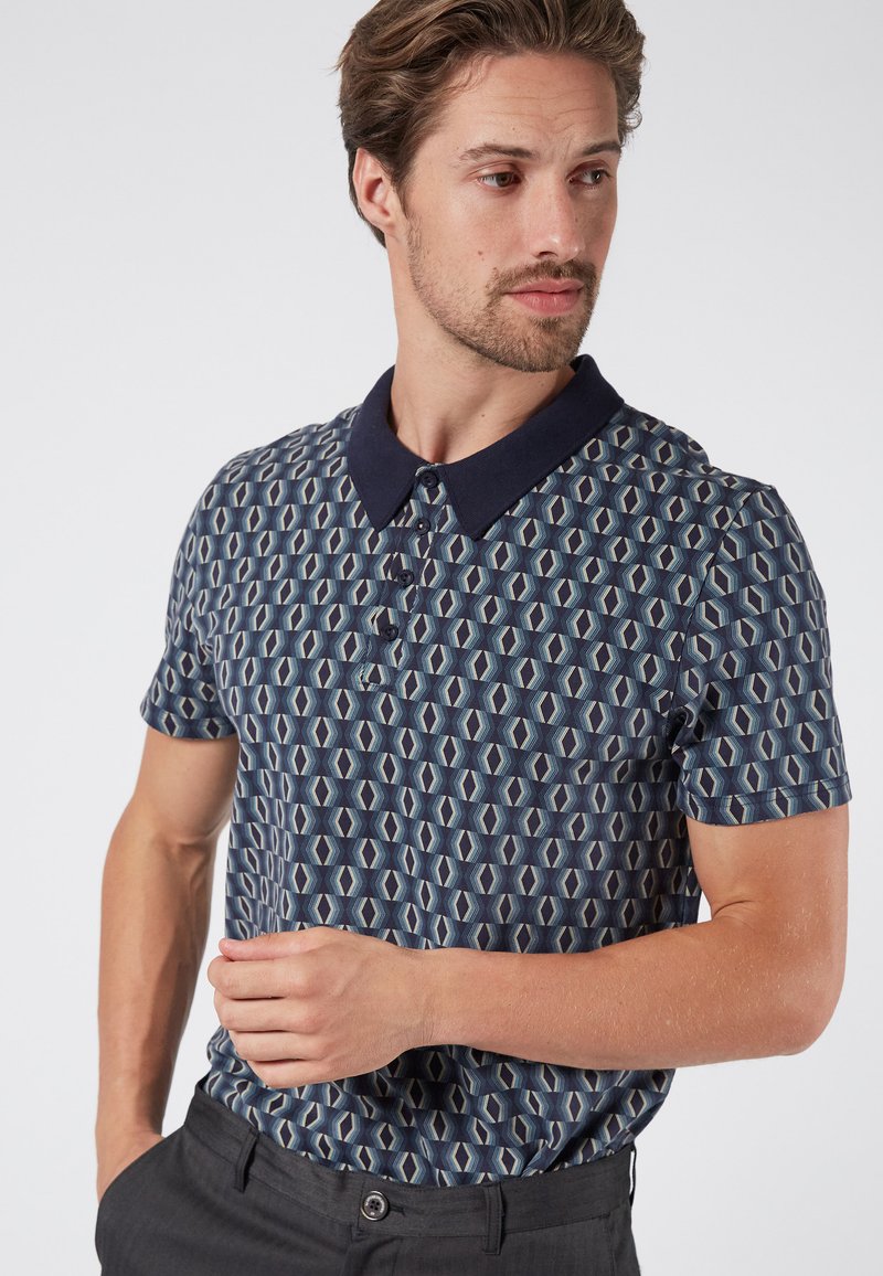 Kortärmad polo skjorta med ett geometriskt mönster i blått och beige, med en marinblå krage och plagg med tre knappar. Slät tygtextur.