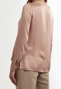 Blusa de seda en un suave tono rosa, con mangas largas con acentos de botones, un escote redondo y una textura suave y ligeramente reflectante.