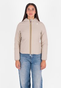 Vrouw in een beige gewatteerde jas met een gestreepte rits aan de voorkant en een blauwe spijkerbroek, staand tegen een witte achtergrond.