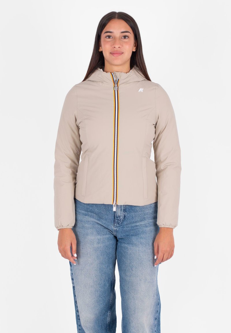 Vrouw in een beige gewatteerde jas met een gestreepte rits aan de voorkant en een blauwe spijkerbroek, staand tegen een witte achtergrond.